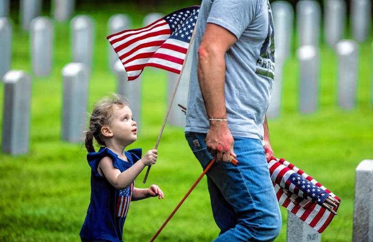 Riley Bell Prince, 3, looks up at a flag as she helps her dad, Matt Prince, plant American flags at the New Hampshire Veterans Cemetery on Thursday, May 23, 2024. Sgt. Prince served in Iraq for eight years and his father served in World War II. Veteran groups and citizens put flags on every marker.