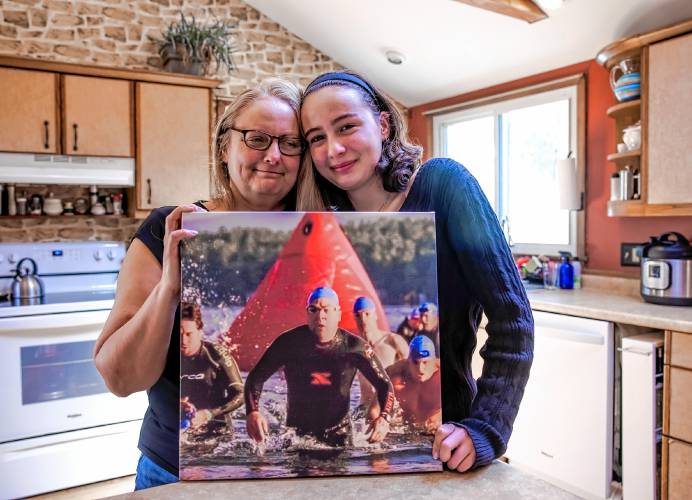 Maia Bradley and her mother, Miiko, hold up a photo of Mike Bradley in one of his triathlon photos in the family kitchen on Saturday, March 30. 2024.