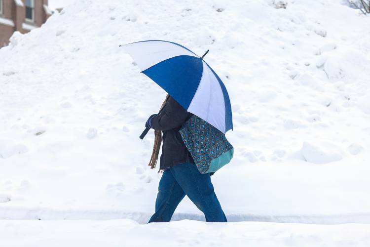 A person with an umbrella walks in front of a pile of snow from the parking lot of Christ The King parking lot on Thursday morning, February 13, 2025.