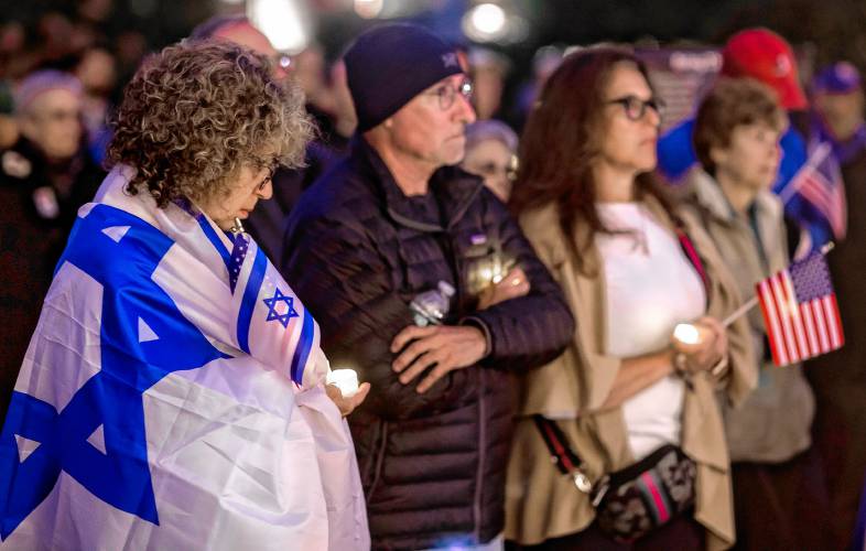 Elise White of Nashua holds a candle as she wraps herself in the Israeli flag during the vigil at the State House on Monday evening, October 7, 2024, to commemorate the first anniversary of the Hamas attack on Israel.